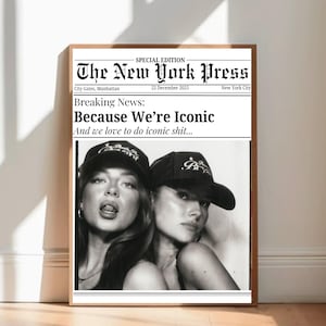 May include: A framed black and white print resembling a newspaper front page. The headline reads "The New York Press" with the date December 22, 2025. The image features two women wearing black caps, with the text "Because We're Iconic".