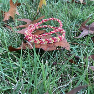 Puede incluir: Una pulsera trenzada en rojo, amarillo y blanco descansa sobre una hoja marrón en medio de hierba verde y hojas caídas. La pulsera está hecha de hilos tejidos apretados, mostrando un patrón colorido.