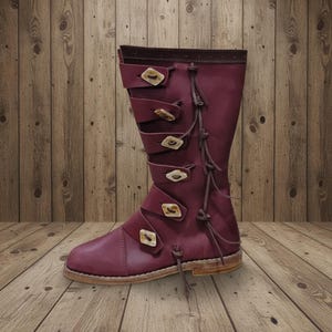 May include: A burgundy leather boot with multiple straps and decorative wooden buckles. The boot has a tall shaft and a rounded toe. Tassels hang from the straps. The boot is displayed on a wooden surface.