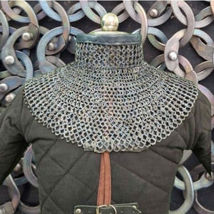 Könnte beinhalten: Nahaufnahme eines mittelalterlichen Kettenhauben, einer schützenden Kopf- und Nackenbedeckung. Die Haube besteht aus kleinen, ineinandergreifenden Metallringen. Sie wird über einem schwarzen, gesteppten Kleidungsstück mit Lederriemen und Schnalle getragen.