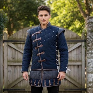 Op de afbeelding: Een marineblauw gewatteerd middeleeuws doublet met bruine leren riemen en accenten. Het kledingstuk heeft een hoge kraag, schoudervullingen en decoratieve witte stiksels langs de zijkanten. De onderste zoom is donkergrijs met decoratieve bruine accenten.