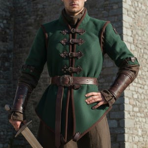 May include: A green medieval-style tunic with brown leather accents, including a belt and decorative straps. The tunic features a high collar and long sleeves with leather cuffs. The person is holding a sword.