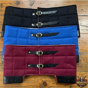 May include: Three padded armor pieces in black, blue, and burgundy. Each piece features a quilted design, black leather straps, and brass buckles. The bottom edges have black leather trim. The items are displayed on a wooden surface.