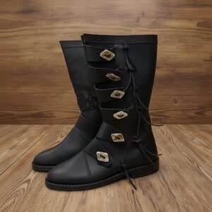 May include: A pair of black leather boots with multiple straps and decorative buttons. The boots have a knee-high design and are set against a wooden background. The boots have a classic, timeless style.