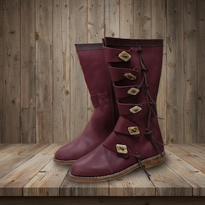 May include: A pair of burgundy leather boots with multiple straps and decorative buttons. The boots have a knee-high design with a side zipper and are displayed on a wooden surface. The boots have a rustic, medieval aesthetic.