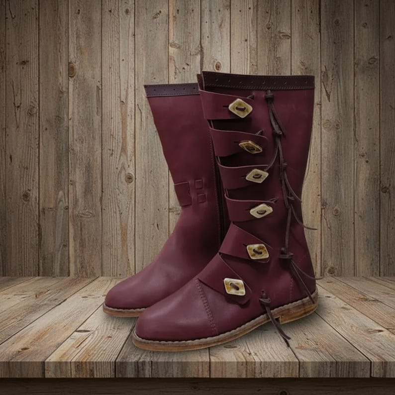 May include: A pair of burgundy leather boots with multiple straps and decorative buttons. The boots have a knee-high design with a side zipper and are displayed on a wooden surface. The boots have a rustic, medieval aesthetic.