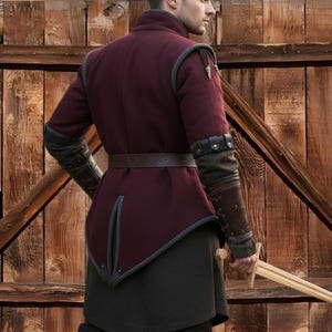 May include: A burgundy medieval-style jacket with gray trim and a brown leather belt. The jacket has long sleeves with dark gray and brown accents. The person is holding a wooden sword. The background is a wooden door.