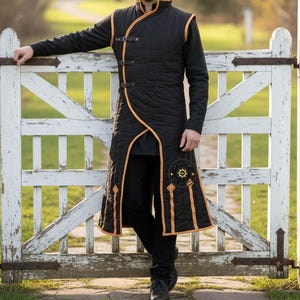 May include: Black quilted vest with gold trim and accents. The vest features a unique asymmetrical design with leather straps and a sun emblem. The garment is worn over a black long-sleeve shirt and pants, creating a layered look.