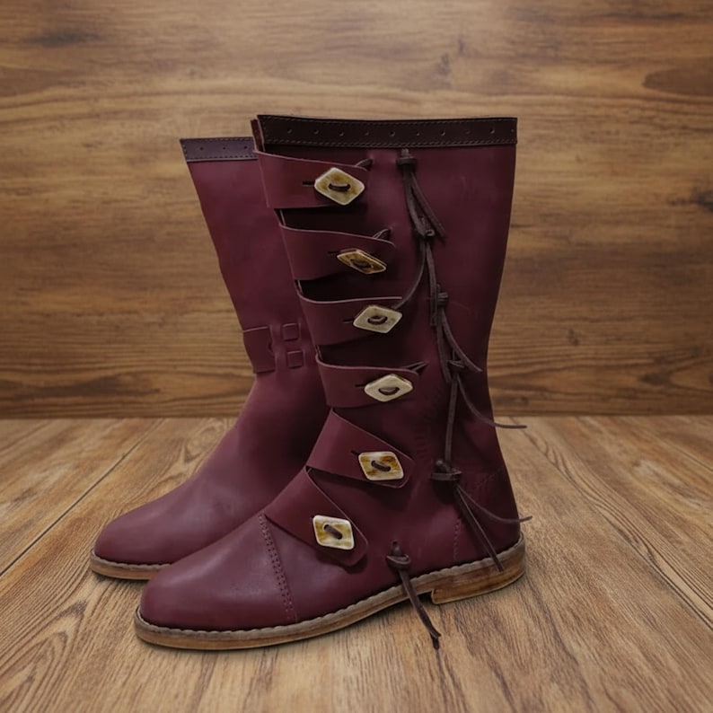 May include: A pair of burgundy leather boots with multiple straps and decorative buckles. The boots have a knee-high design with a rounded toe and are displayed on a wooden surface. The buckles are a light tan color.