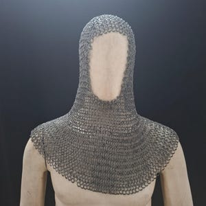 May include: A chainmail coif, a medieval head covering, is displayed. The coif is made of small, interlocking metal rings, creating a flexible, protective hood. The coif covers the head, neck, and shoulders, with a face opening.