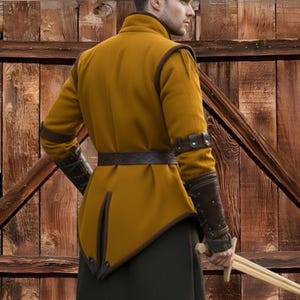 May include: Mustard-colored medieval tunic with brown leather accents. The tunic features a high collar, long sleeves, and a decorative belt. Brown leather arm guards are also visible. The tunic is worn over dark green pants. A wooden sword is held.