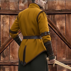 May include: Mustard-colored medieval tunic with brown leather accents. The tunic features a high collar, long sleeves, and a decorative belt. Brown leather arm guards are also visible. The tunic is worn over dark green pants.