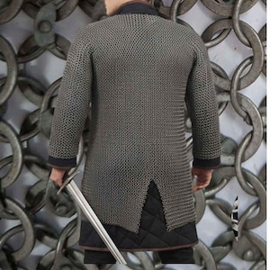 May include: A silver chainmail shirt with long sleeves and a split hem. The shirt has a black collar and cuffs. A person is holding a silver sword. The background features a large chain design.