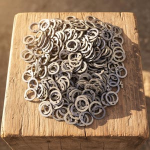 May include: A collection of small, silver metal washers, each with a circular form and a small hole. They are scattered on a rough wooden surface, possibly a block or table. The washers are made of metal.