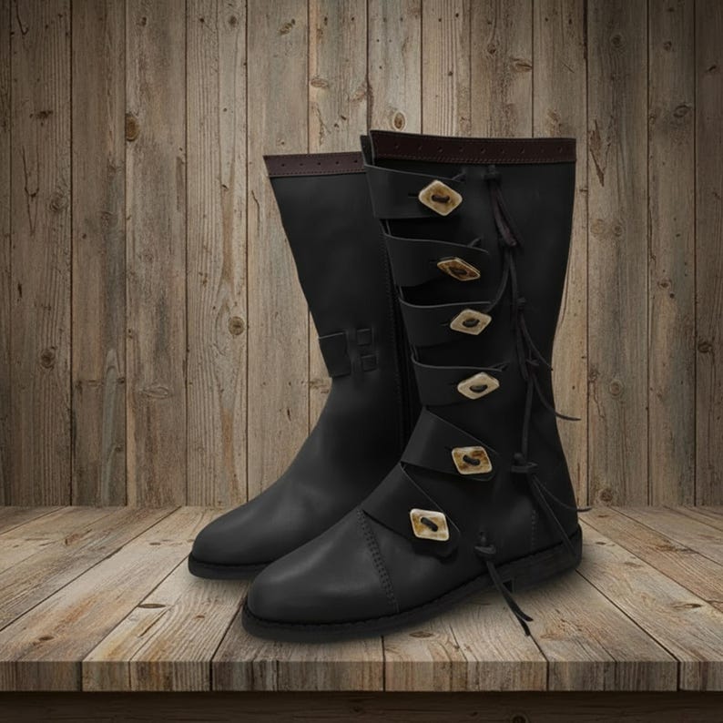 May include: A pair of black leather boots with decorative straps and buckles. The boots have a knee-high design with brown accents at the top and feature multiple straps with decorative buckles. The boots are displayed on a wooden surface.