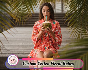 May include: A woman wearing a custom cotton floral robe with a vibrant red and floral pattern. She is sitting outdoors and drinking from a coconut. The robe is tied at the waist. The text "Custom Cotton Floral Robes" is visible.
