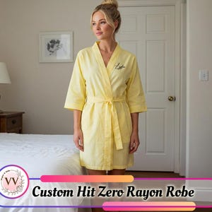 Peut inclure: Un peignoir en rayonne jaune avec une ceinture assortie, porté par une personne. Le peignoir a des manches trois-quarts et un nom personnalisé brodé sur le devant. Le texte "Custom Hit Zero Rayon Robe" est affiché en bas de l'image.