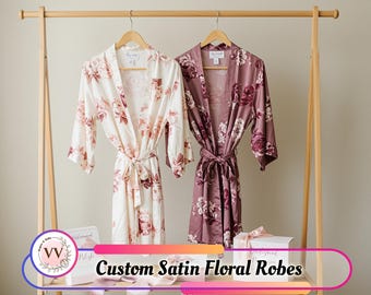May include: Two satin floral robes hang on a wooden rack. One is cream-colored with pink flowers, the other is a mauve color with darker pink flowers. Both robes have a matching tie belt. The text "Custom Satin Floral Robes" is visible.