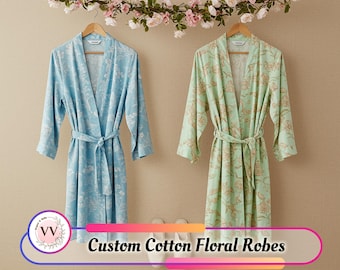 May include: Two custom cotton floral robes, one light blue and one mint green, hang on wooden hangers. The light blue robe has white floral patterns, and the mint green robe has beige floral patterns. The text "Custom Cotton Floral Robes" is displayed.