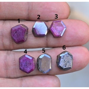 Puede incluir: Seis piedras preciosas de rubí en bruto de varios tonos de rojo y morado. Las piedras son hexagonales o de forma irregular, con texturas naturales visibles. Las gemas se muestran sobre un fondo neutro.