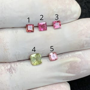 Puede incluir: Se muestran cinco piedras preciosas de varios colores y formas. Tres son de corte cuadrado, con tonos rosados ​​y rojos. Una es un cuadrado verde claro, y la última es una gema rosa, con corte cojín. Las gemas están sobre un fondo blanco.