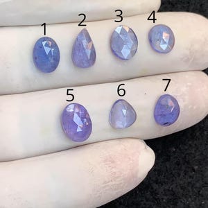 Puede incluir: Se muestran siete piedras preciosas de tanzanita facetadas, de forma ovalada y de lágrima, en varios tonos de azul y púrpura. Las piedras preciosas están dispuestas sobre una superficie blanca, mostrando sus diferentes cortes y colores. Las piedras preciosas están numeradas del 1 al 7.