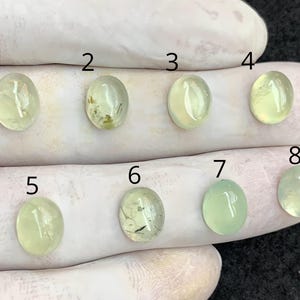 May include: Eight oval-shaped, translucent gemstones in varying shades of pale green and yellow are displayed. The gemstones are numbered 1 through 8. The stones are set against a white background, likely a gloved hand.