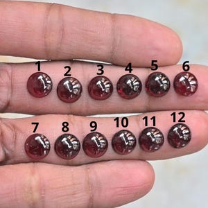 Op de afbeelding: Twaalf ronde, gepolijste, donkerrode edelstenen worden getoond. Elke edelsteen heeft een licht gewelfd oppervlak en reflecteert licht. De edelstenen zijn gerangschikt in drie rijen van vier, genummerd van 1 tot 12.
