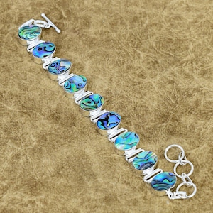 Puede incluir: Pulsera de plata con conchas de abulón ovaladas en tonos azules, verdes y morados. La pulsera tiene un cierre de palanca. Las conchas de abulón están engastadas en biseles de plata. Sobre una superficie marrón.