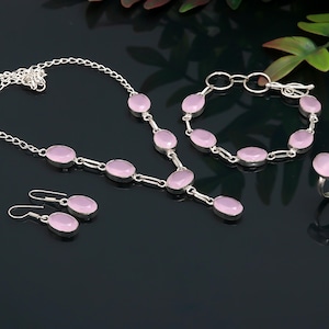 May include: A jewelry set featuring a necklace, bracelet, earrings, and ring. Each piece has a silver-colored metal setting and oval, light pink gemstones. The necklace and bracelet are linked with small chain segments. The earrings are drop style.