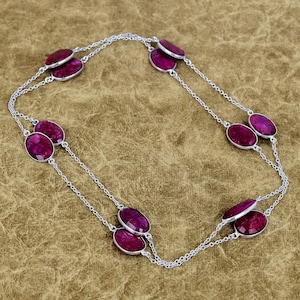 May include: A silver necklace featuring oval-shaped, deep red gemstones. The gemstones are set in silver bezels and connected by delicate silver chains. The necklace is displayed against a neutral background, highlighting the jewelry's design and color.