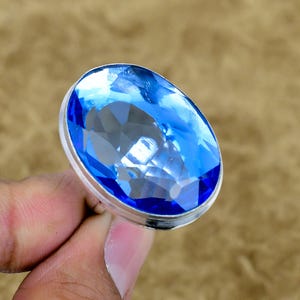 Puede incluir: Un anillo con una gran piedra preciosa azul de forma ovalada y tallada, engastada en una banda de metal de tono plateado pulido. La piedra muestra un color azul vibrante y translúcido con múltiples facetas geométricas.
