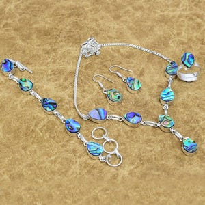 May include: A jewelry set featuring a necklace, bracelet, earrings, and ring. The set is made of silver and features iridescent abalone shell teardrop-shaped stones in shades of blue, green, and purple. The ring has a single abalone stone.