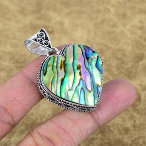 May include: A heart-shaped pendant with a silver-colored bail and a vibrant abalone shell inlay. The shell displays iridescent colors of green, blue, and gold. The pendant is held by a hand.