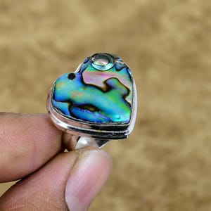 Può includere: Un anello a forma di cuore con una fascia argentata e un vivace elemento centrale in conchiglia di abalone. Il guscio mostra blu, verdi iridescenti e sfumature di viola. L'anello è tenuto dalle dita di una persona.