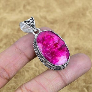 May include: A silver pendant with an oval, vibrant pink gemstone. The gemstone is set in a detailed, ornate silver frame. The pendant has a decorative bail at the top for a chain. The design is intricate and eye-catching.