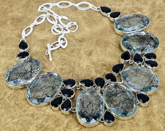 May include: A silver necklace with oval, clear gemstones with black inclusions, and black teardrop-shaped stones. The necklace has a silver chain and clasp. The background is brown.