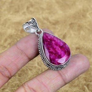 May include: A teardrop-shaped pendant with a large, faceted, magenta gemstone. The gemstone is set in a silver-colored metal frame with intricate detailing. The pendant has a decorative bail for attaching to a chain.