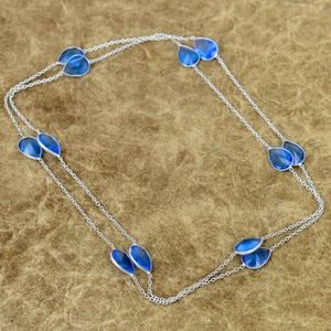 Può includere: Una lunga collana a catena argentata con molteplici pietre traslucide blu a forma di goccia, distanziate uniformemente lungo le maglie. La collana è disposta in forma rettangolare, mettendo in risalto la catena metallica e gli accenti blu.
