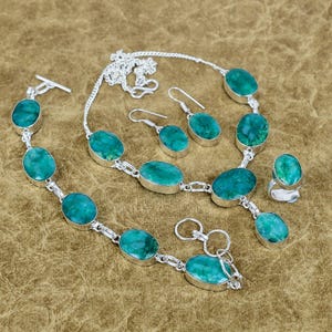 Puede incluir: Un conjunto de joyas que incluye un collar, una pulsera, pendientes y un anillo. Cada pieza tiene piedras ovaladas de color turquesa engastadas en metal plateado. El conjunto se muestra sobre una superficie marrón.