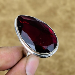 Può includere: Anello con una grande pietra preziosa rosso scuro a forma di goccia, sfaccettata, con una fascia argentata. La pietra è il punto focale, che mostra il suo colore ricco e le sue intricate sfaccettature. Sfondo neutro.