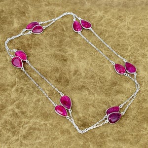 May include: A silver necklace featuring multiple teardrop-shaped, magenta-colored gemstones. The gemstones are set in silver and connected by delicate silver chains. The necklace is displayed on a brown surface.