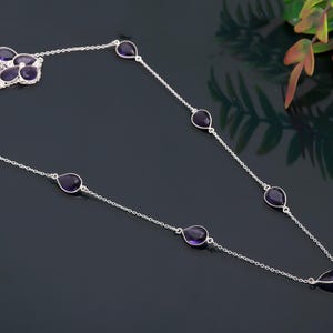 May include: A silver necklace with a floral cluster and teardrop-shaped purple gemstones. The necklace has a delicate chain design with evenly spaced gemstones. The gemstones are a deep purple colour, and the necklace is displayed on a dark surface.