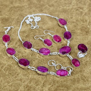 May include: A jewelry set featuring a necklace, bracelet, earrings, and ring. Each piece has oval-shaped, deep pink gemstones set in silver-colored metal. The necklace and bracelet have a chain-link design, and the earrings are simple drop style.
