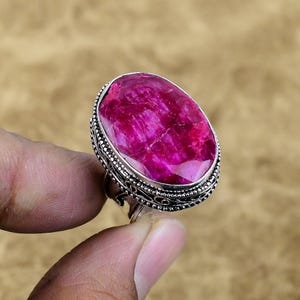 Puede incluir: Un anillo de plata con una gran gema de rubí de corte ovalado. El rubí es de un tono rojo-rosa vibrante, con patrones internos visibles. El engaste del anillo presenta un diseño ornamentado y detallado alrededor de la gema.