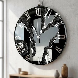 Könnte beinhalten: Eine runde, schwarze Wanduhr mit silbernen römischen Ziffern und Zeigern. Das Zifferblatt hat ein einzigartiges Design mit strukturierten, dunkelgrauen und weißen Akzenten. Die Uhr hängt an einer grauen Wand.