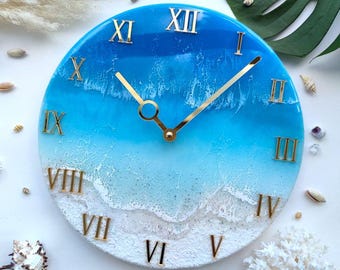 Puede incluir: Reloj de pared redondo con diseño de olas oceánicas azules y blancas. La esfera del reloj presenta números romanos dorados y manecillas doradas. Es una pieza decorativa con temática costera, de unos 25 cm de diámetro.