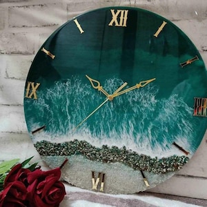 Puede incluir: Un reloj de pared redondo con un diseño de olas oceánicas turquesas y blancas. La esfera del reloj presenta números romanos y manecillas doradas. La parte inferior del reloj tiene un diseño de playa arenosa con pequeñas piedras.