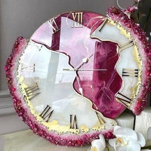 Op de afbeelding: Een decoratieve wandklok met een uniek ontwerp. De wijzerplaat heeft een mix van wit en kastanjebruin hars met gouden Romeinse cijfers. De buitenrand is versierd met roze en rode kristallen, wat een opvallend visueel effect creëert.
