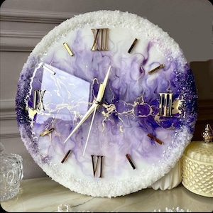 May include: A circular wall clock with a white, purple, and gold design. The clock face has Roman numerals and gold hands. The clock's edge is decorated with a white, textured border. A decorative home decor item.